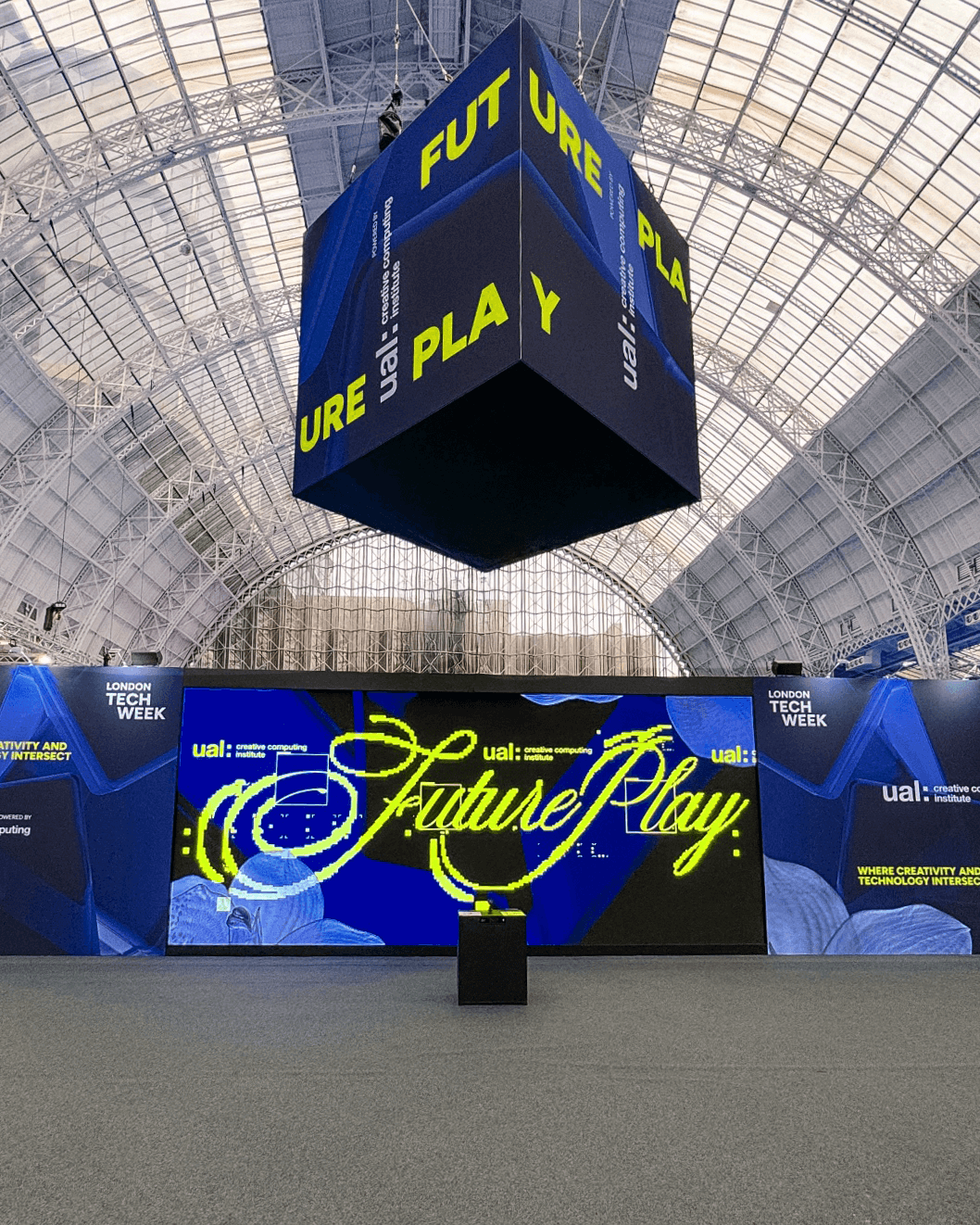Exhibition space for London Tech Week with large hanging cube and digital screen displaying 'Future Play' and 'ual: creative computing institute' in a glass-roofed venue.