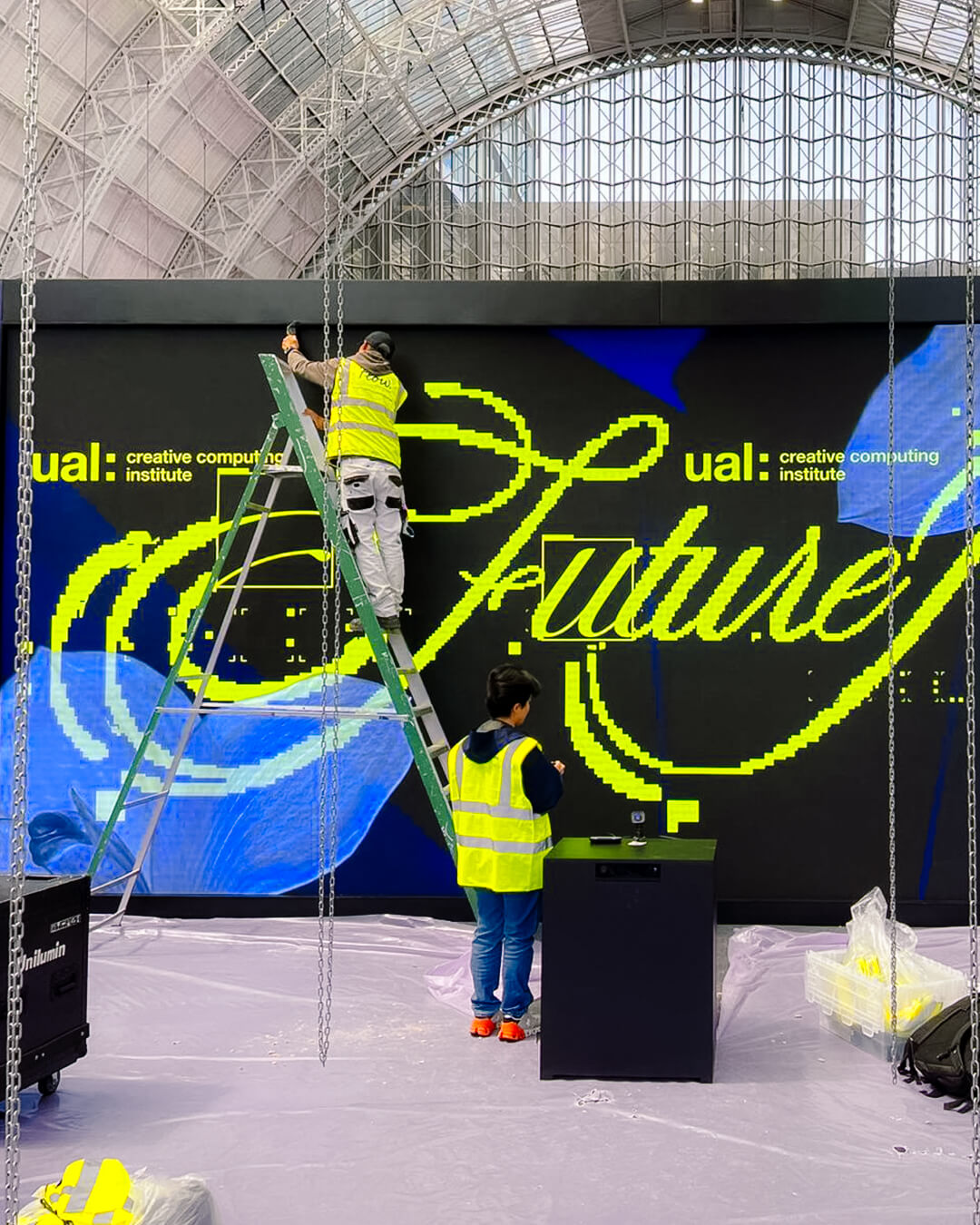 Two workers in yellow safety vests install a large digital screen displaying 'ual: creative computing institute' and the word 'future' in bright yellow script at an indoor venue.
