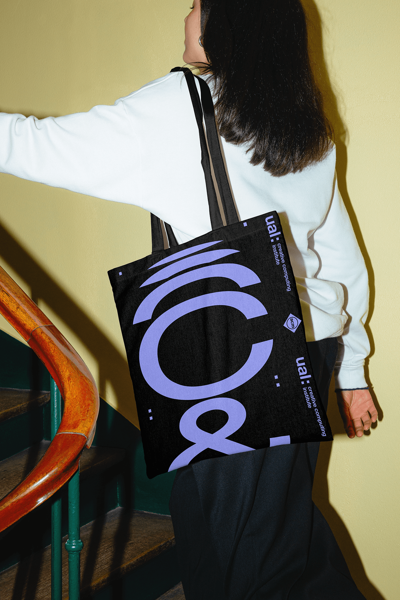 Person carrying a black tote bag with large purple text on the strap of a staircase with wooden railing and green banister.