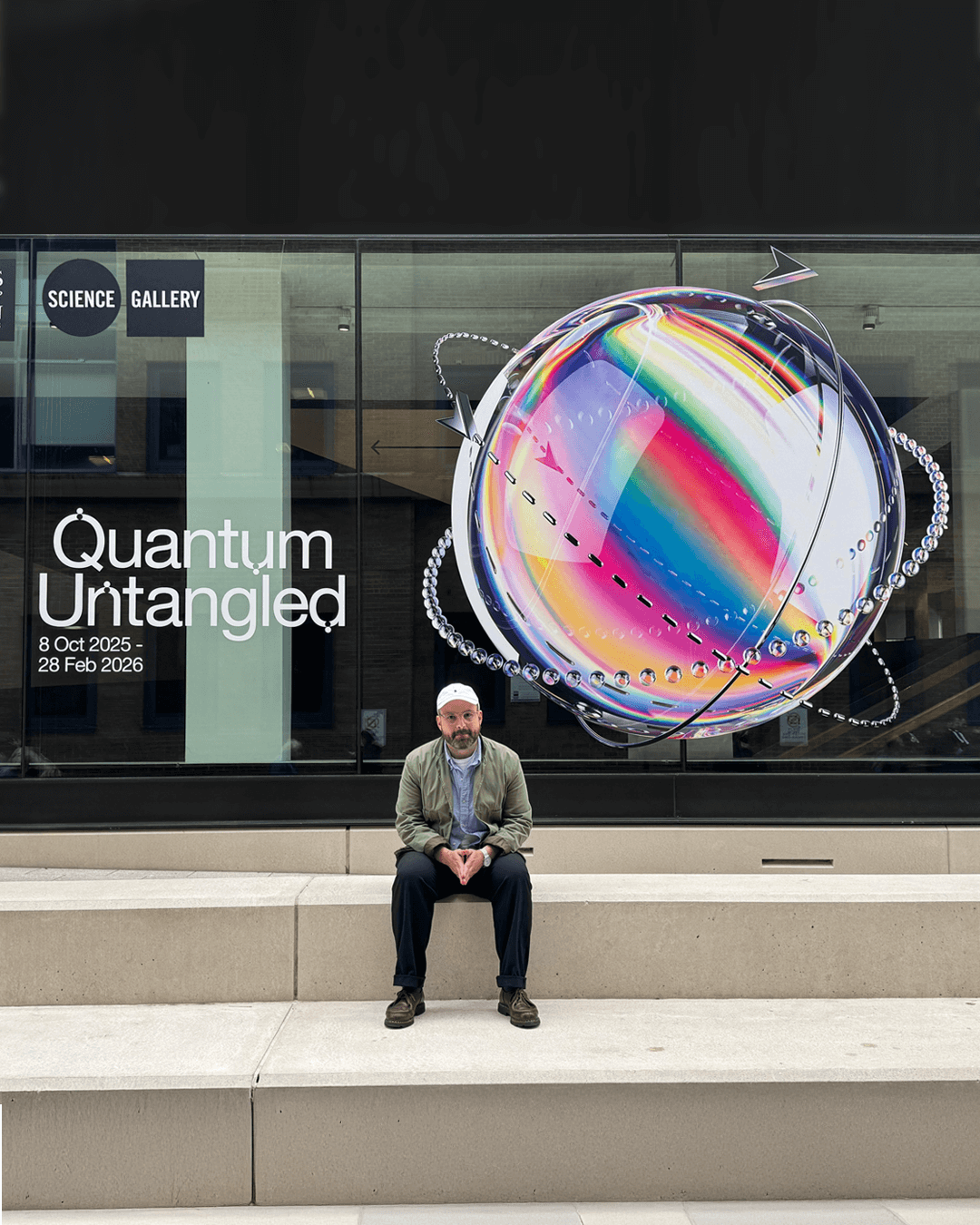 Man sitting on beige stone steps in front of a glass wall displaying a colorful spherical graphic and text reading Quantum Untangled, 8 Oct 2025 - 28 Feb 2026 at Science Gallery.