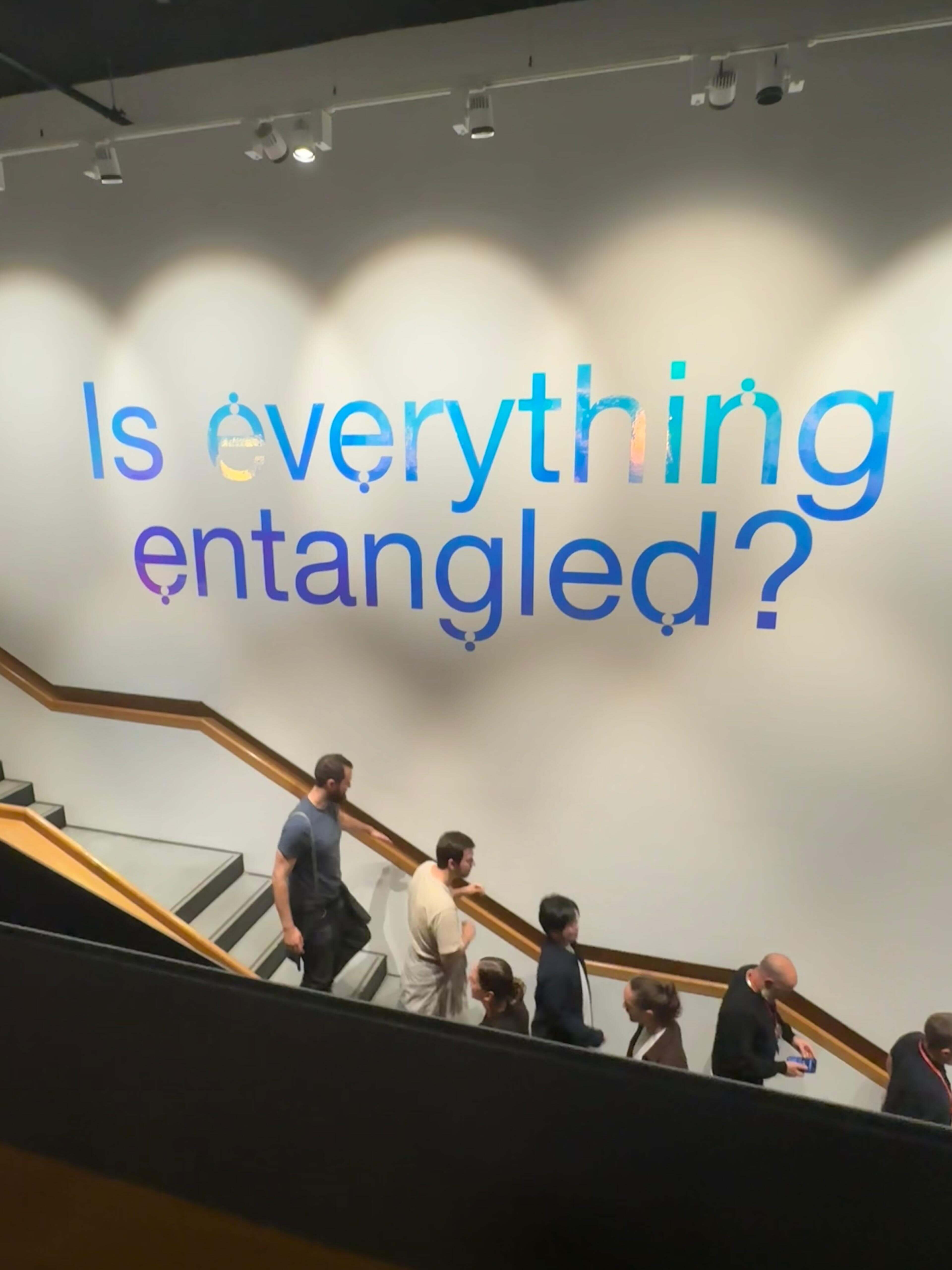 People walking down a staircase with a wall sign reading 'Is everything entangled?' in large blue letters.