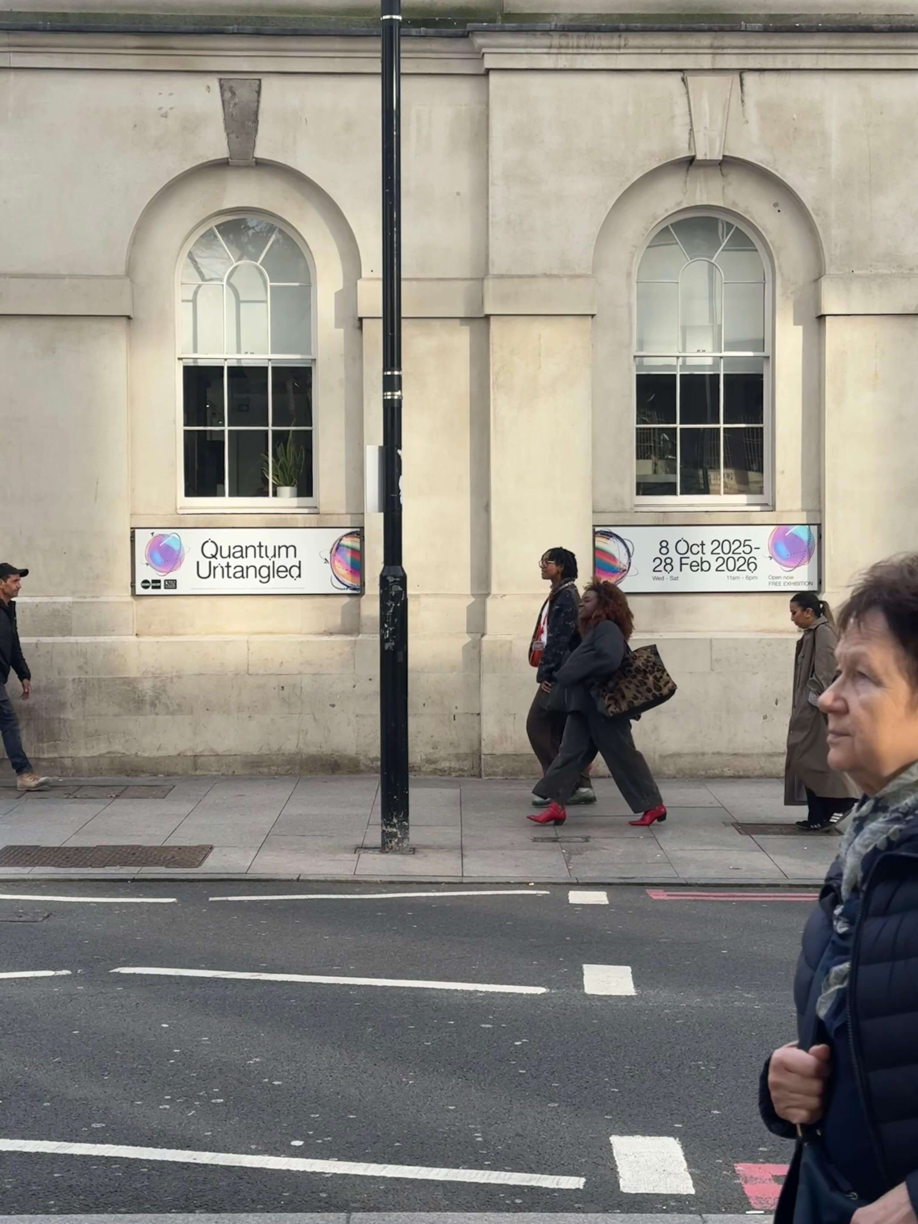 People walking past a beige building with two arched windows displaying signs for Quantum Untangled exhibition dated 8 Oct 2025 to 28 Feb 2026.