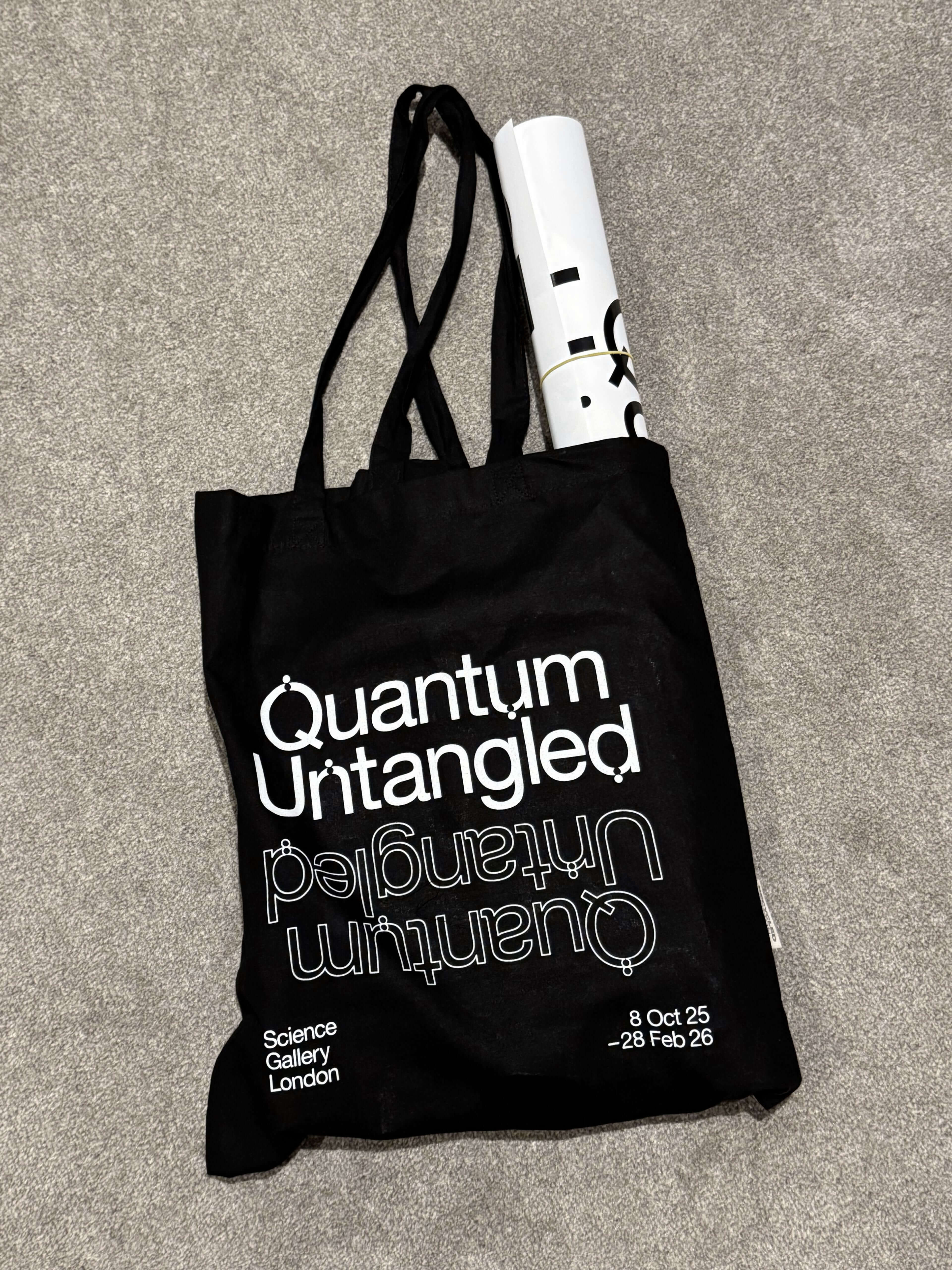 Black tote bag with white text reading 'Quantum Untangled' twice, along with 'Science Gallery London' and exhibition dates '8 Oct 25 - 28 Feb 26', lying on a gray carpet with a rolled-up poster inside.