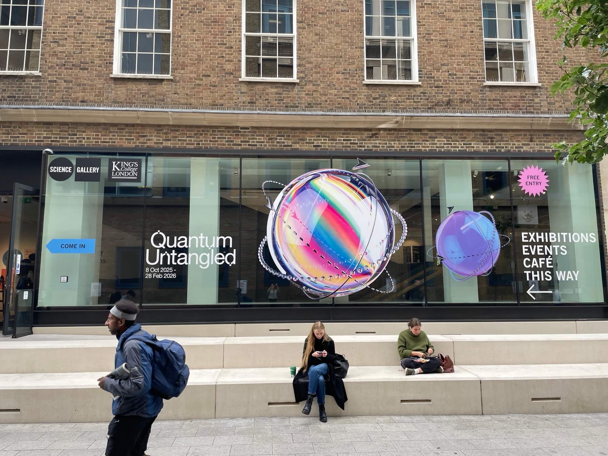 Exterior window display of King's College London Science Gallery advertising the Quantum Untangled exhibition from Oct 8, 2025 to Feb 28, 2026, with colorful orbiting spheres and signs directing to exhibitions, events, and cafe.