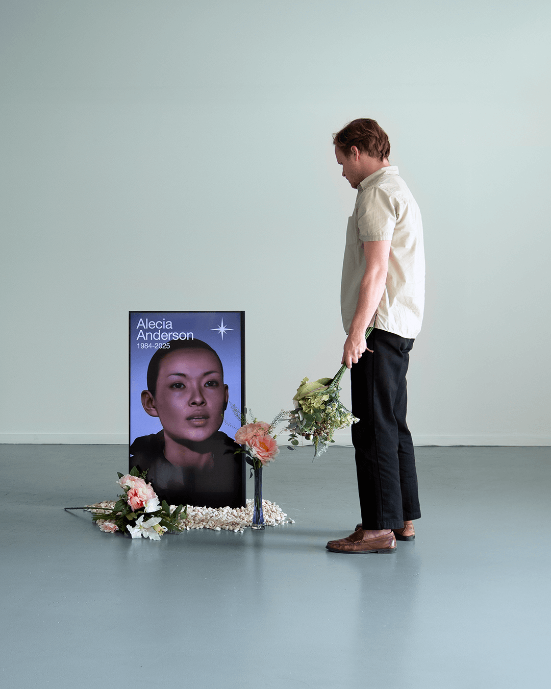 Man in beige shirt and black pants standing with bouquet of flowers, looking at a memorial poster with a woman's face and the text 'Alecia Anderson 1984-2025' surrounded by flowers.