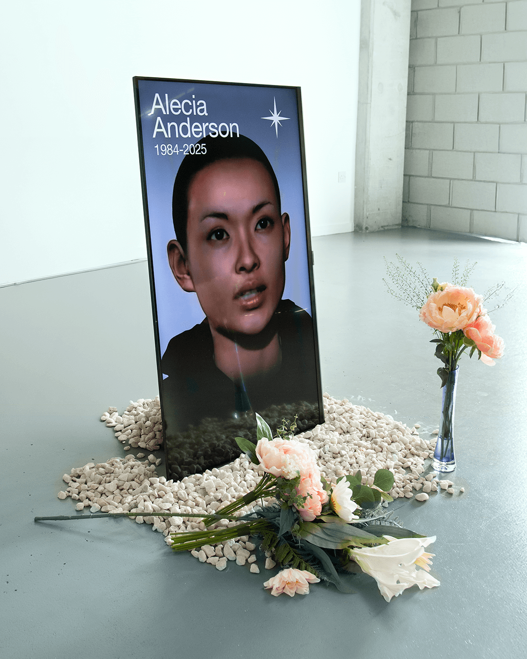 Memorial setup with a digital portrait of Alecia Anderson (1984-2025) surrounded by white pebbles and pink and white flowers on a gray floor.
