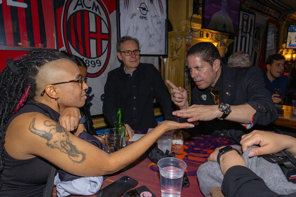 Group of people at a bar table, one man gesturing while a woman with a tattoo holds out her hand.
