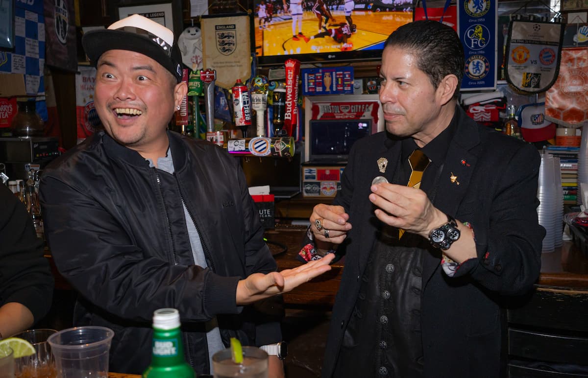 Two men at a bar, one in a black jacket and cap with a surprised expression, the other holding a coin.