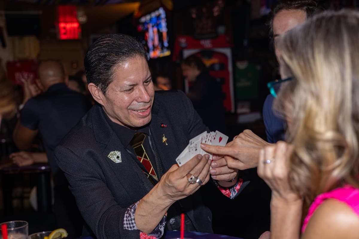 Man smiling and performing a card trick to another person in a bar setting.