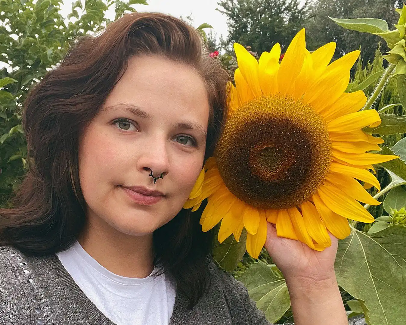 Woman with dark hair holding a large sunflower beside her face in a garden.