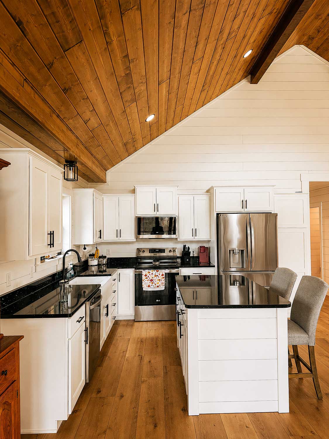 Bright custom kitchen with white shaker cabinetry, black quartz countertops, stainless steel appliances, a center island, and warm tongue-and-groove wood ceilings.
