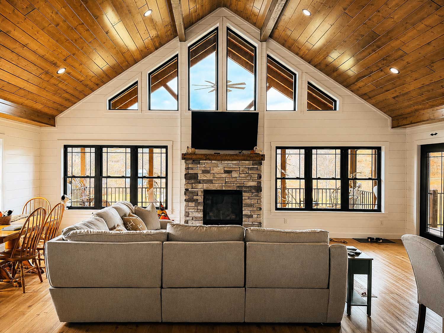 Expansive great room featuring soaring cathedral ceilings, exposed timber beams, a floor-to-ceiling stone fireplace, oversized windows, and an open-concept layout filled with natural light.