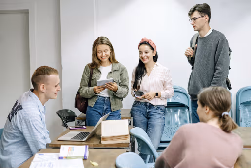 Five young adults in a classroom, two seated at a table and three standing, smiling and chatting while holding notebooks and phones.