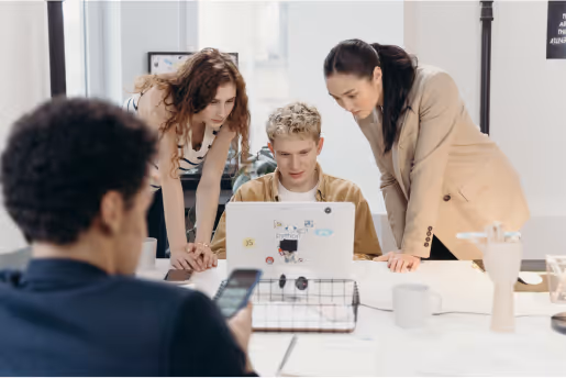 Three colleagues standing around a seated coworker looking at a laptop, while another person in the foreground checks their phone in a modern office setting.