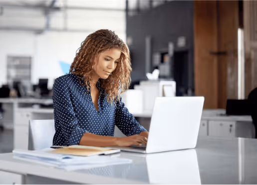 Young woman with curly hair working on a laptop at a desk in a modern, bright office, with papers and a notebook in front of her.