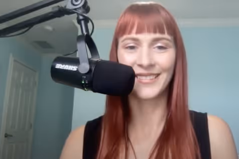 Smiling woman with long red hair speaking into a black Shure microphone in a home recording setup.