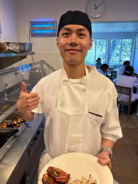 Chef in white uniform and black hat giving thumbs up while holding a plate of food in a kitchen.
