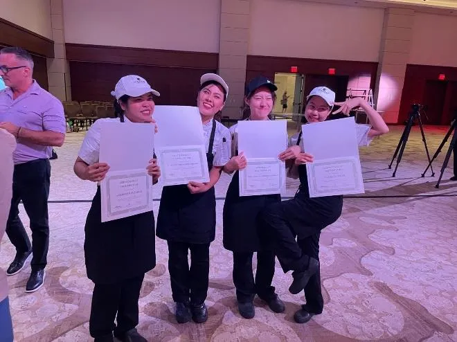 Four people in uniforms holding certificates and smiling indoors on carpeted floor.