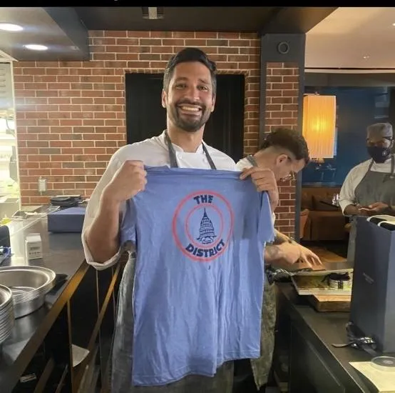 Man smiling and holding a light blue T-shirt with a Capitol dome logo and text 'The District'.