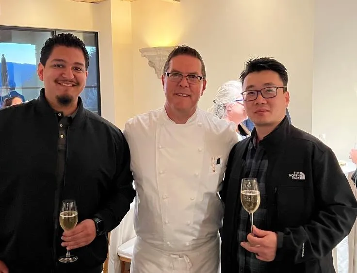 Three men standing indoors, two holding champagne glasses, one wearing a chef's white uniform.
