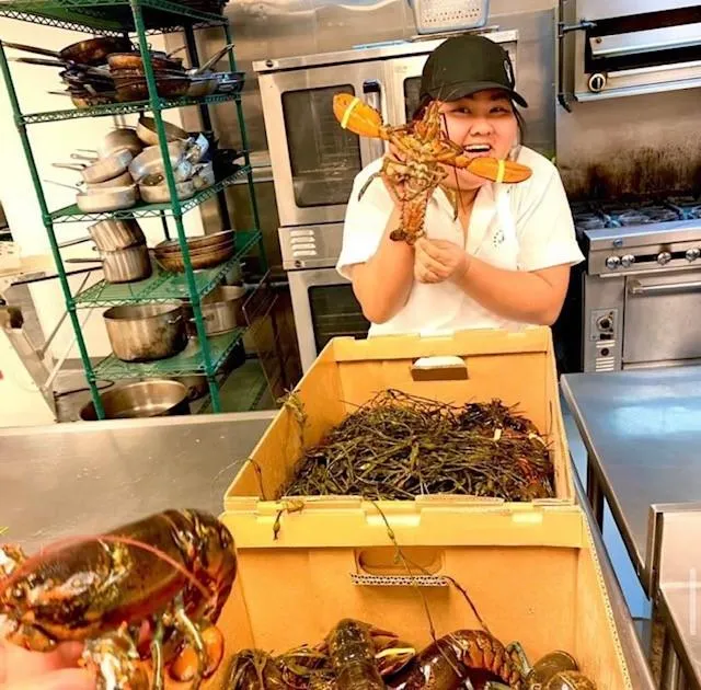 Woman in kitchen holding a lobster above a crate filled with lobsters and seaweed.