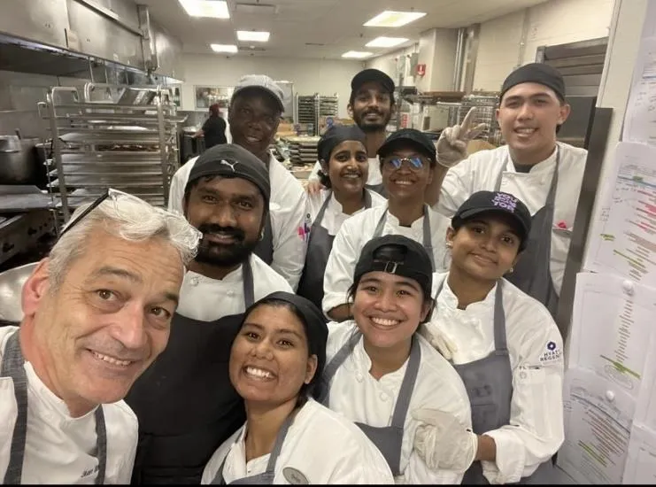 Group of smiling diverse chefs in a kitchen posing for a selfie, wearing white uniforms and aprons.