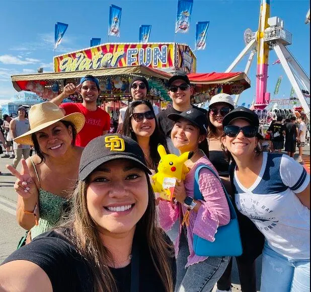 Group of eight smiling people taking a selfie at an outdoor amusement park on a sunny day.
