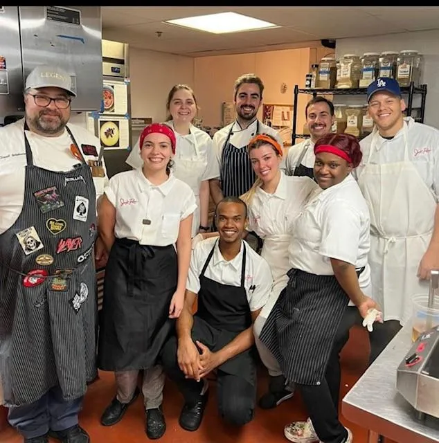 Group of eight smiling kitchen staff wearing aprons and chef uniforms posing in a commercial kitchen.