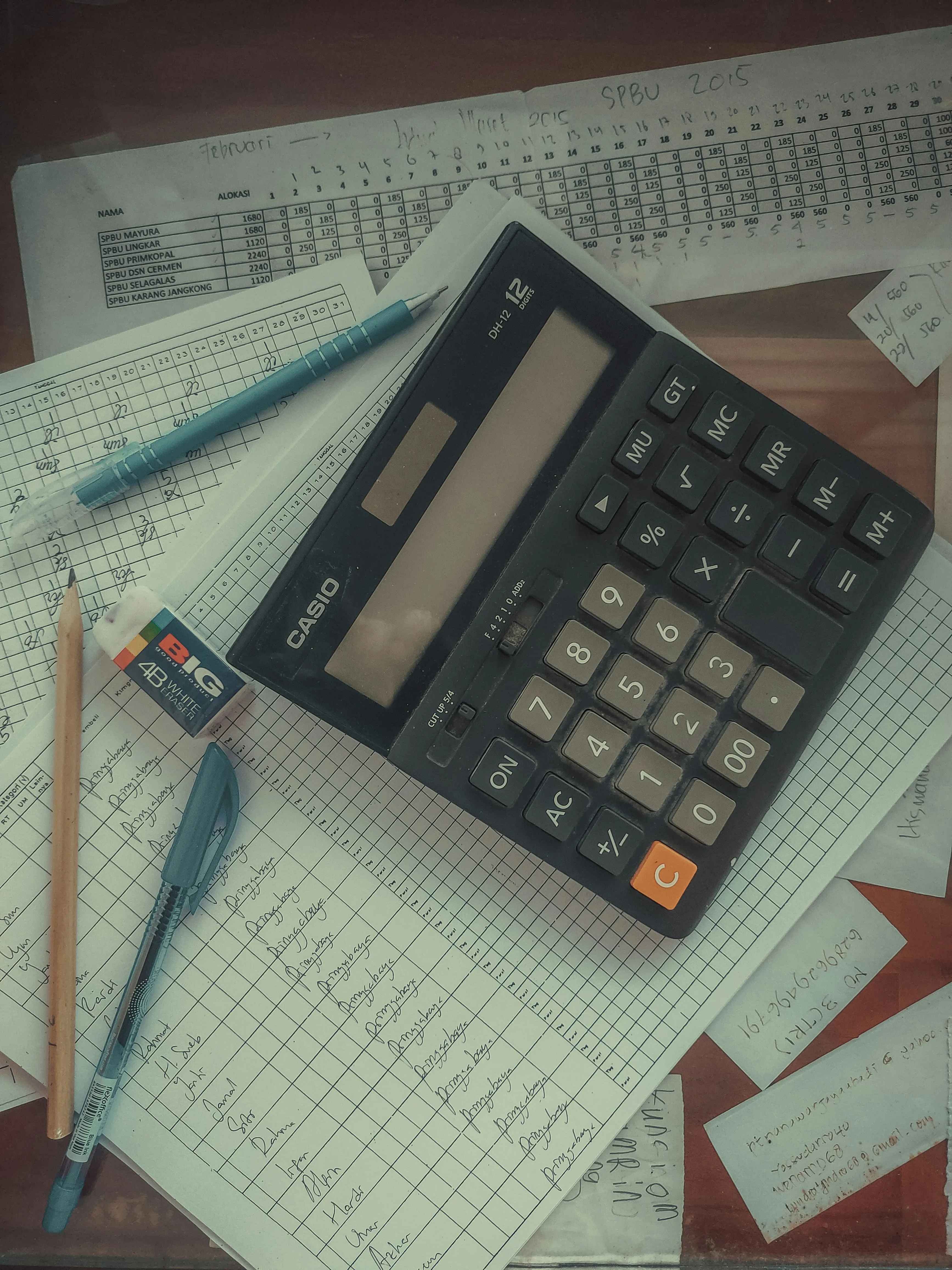 Calculator, pen, pencil, eraser, and handwritten ledger sheets on a wooden desk.
