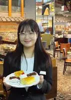 Woman with glasses and black jacket holding a white plate with breakfast foods in a restaurant.