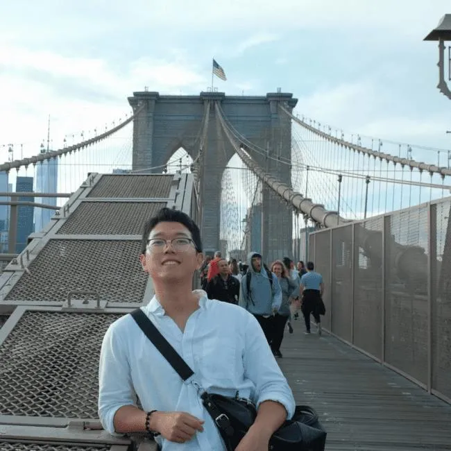 Young man wearing glasses and white shirt posing on Brooklyn Bridge with people walking behind him.