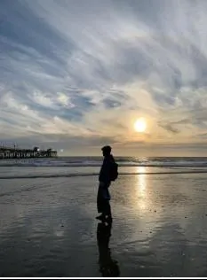 Silhouette of a person standing on wet sand at the beach during sunset with a pier in the background.