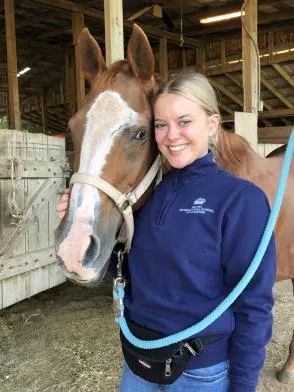 Smiling woman in blue sweatshirt hugging a brown horse with a white blaze in a stable.