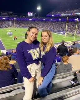 Two women smiling in purple University of Washington shirts at a crowded football stadium.