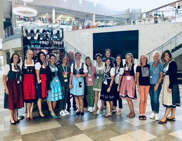 Group of adults standing indoors, many wearing traditional Bavarian dresses, posing and smiling.