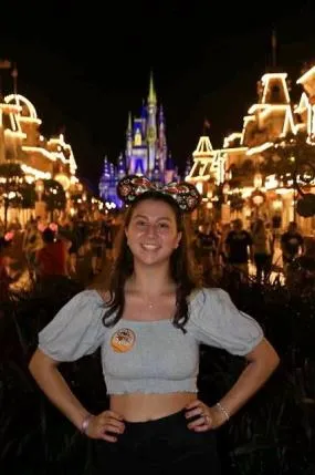 Smiling woman wearing Minnie Mouse ears and crop top standing on a lit street with Cinderella’s Castle in background.