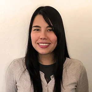 Smiling woman with long dark hair wearing a light gray cardigan and dark shirt against a plain wall.