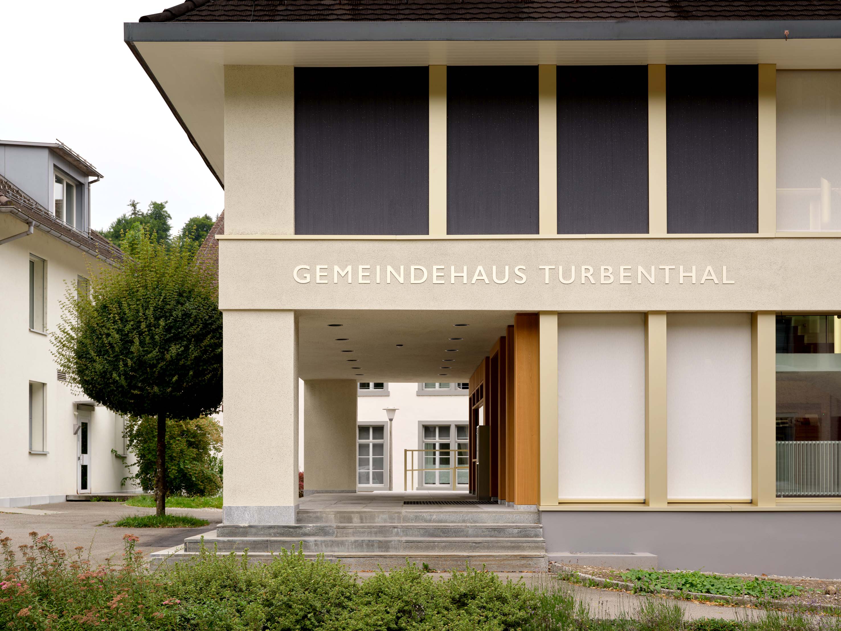 Überdachter Zugang zum Gemeindehaus Turbenthal mit Steinstufen, Stützen und Fassadenbeschriftung.