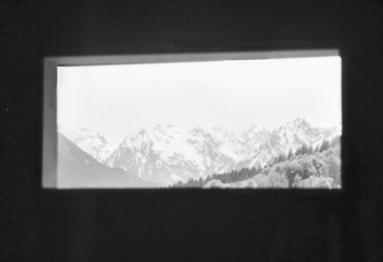 Blick durch das breite Fenster von Haus Conters auf schneebedeckte Berggipfel.