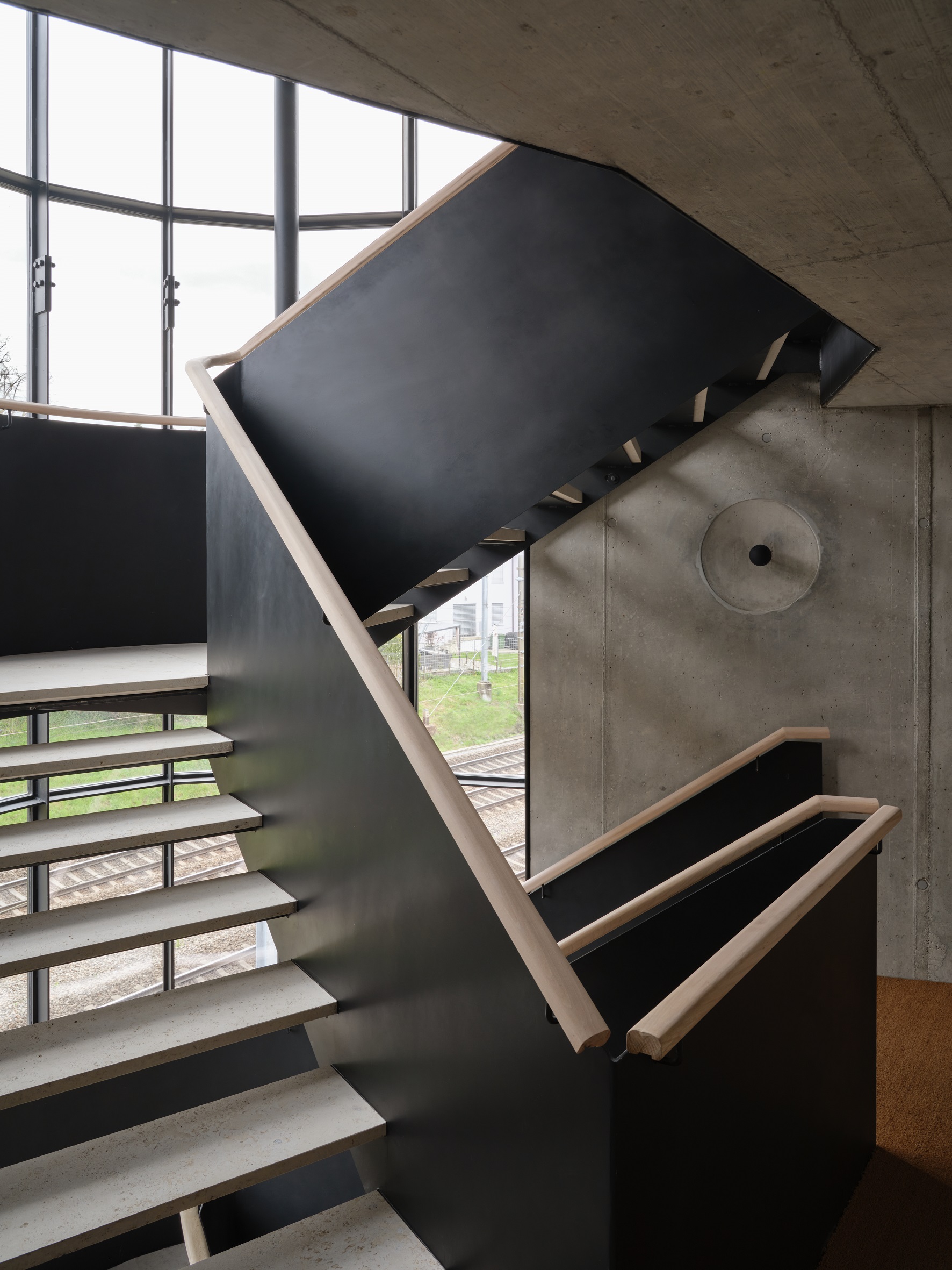 Treppenhaus im Ergänzungsbau Lindeneck mit Sichtbeton, dunkler Stahlbrüstung und Holzhandlauf.