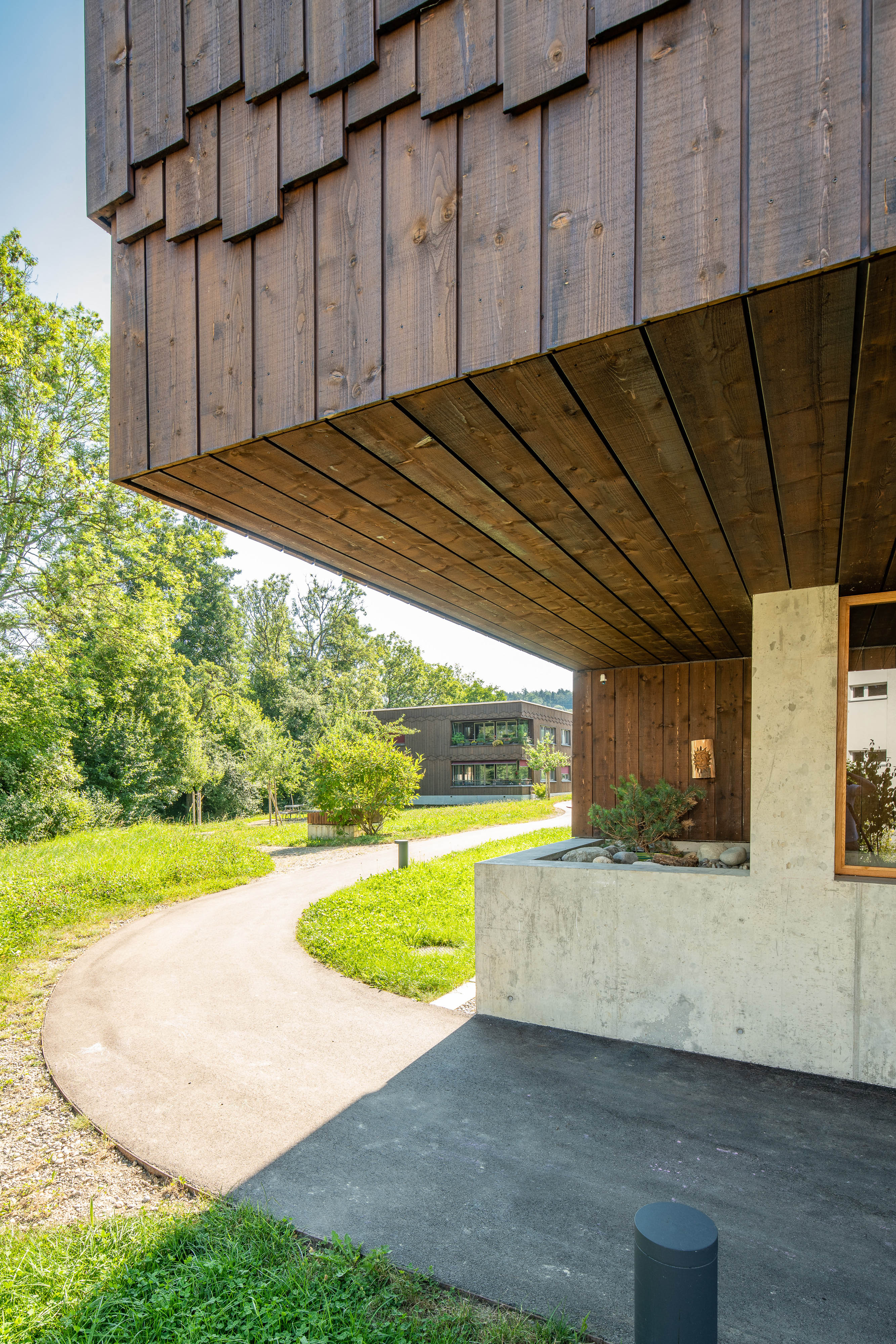 Geschwungener Weg am Eingang der Wohnanlage Grünmatt unter der auskragenden Holzfassade.