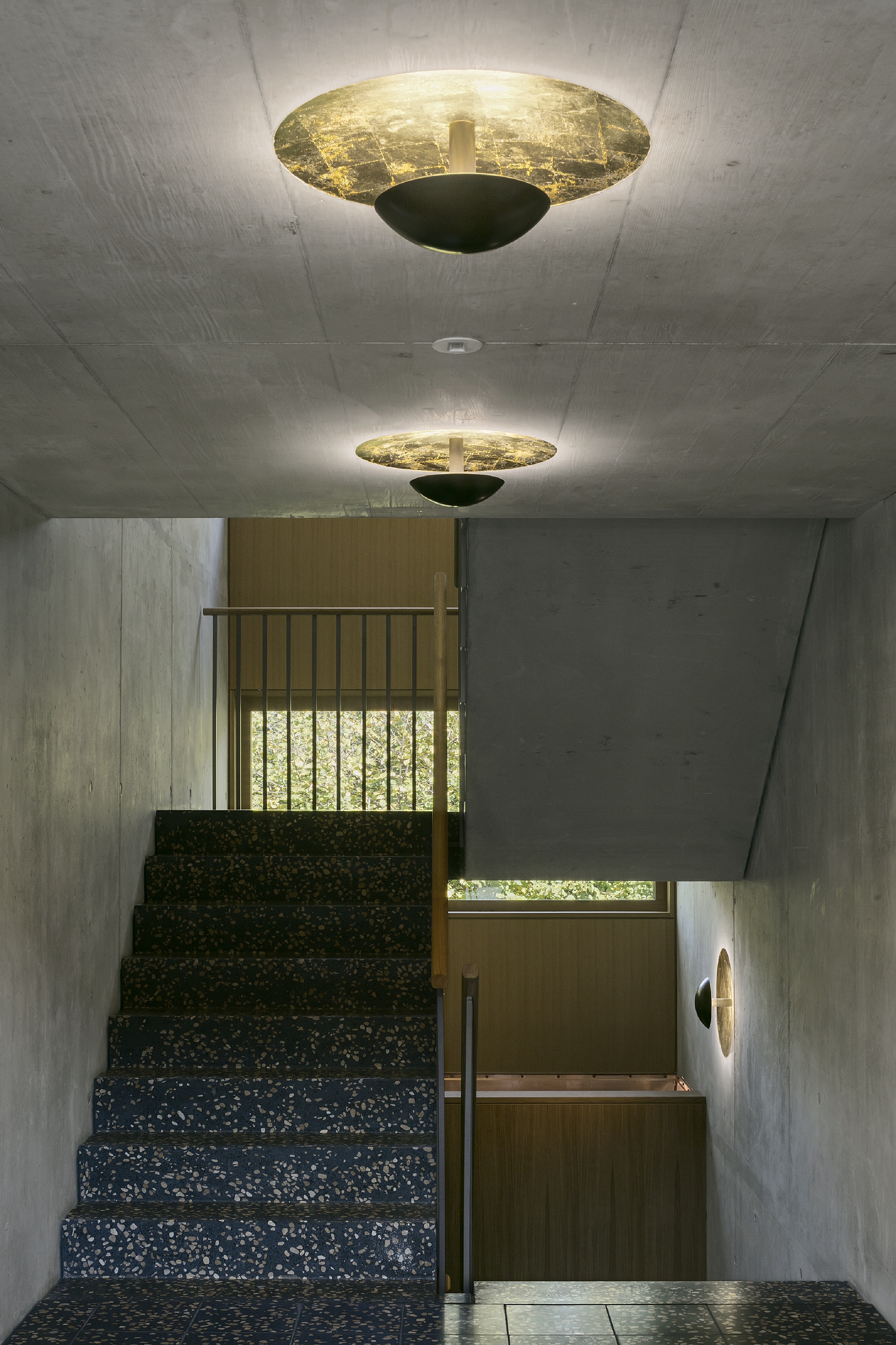 Treppenhaus in Haus B der Wohnanlage Grünmatt mit Sichtbetonwänden und goldfarbenen Deckenleuchten.