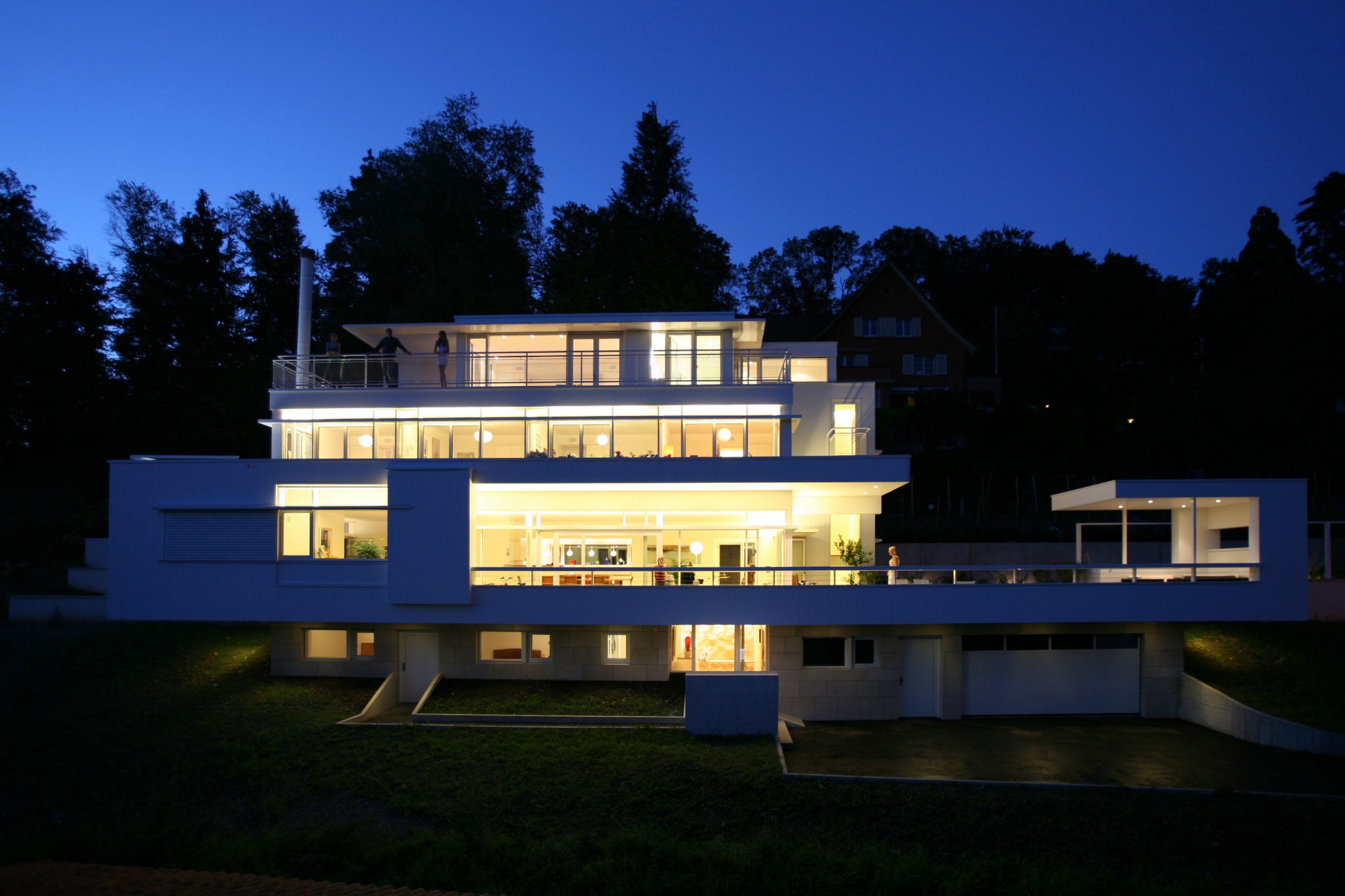 Beleuchtete Südfassade von Haus Trottenweg mit Balkonen und grossen Fensterflächen bei Nacht.
