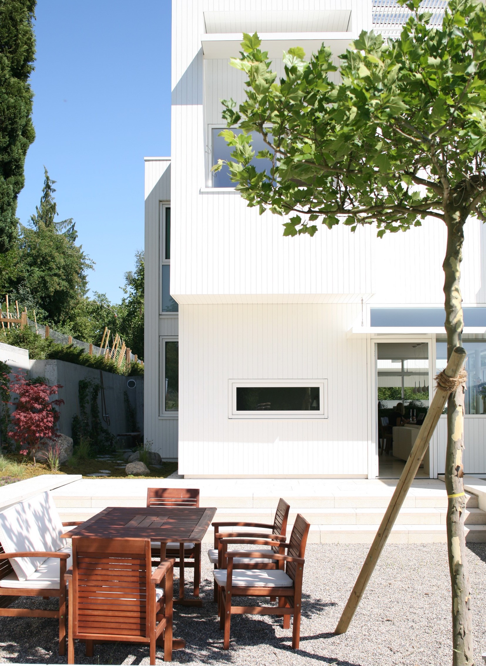 Gartenterrasse mit Holztisch und Baum vor einer weiss verkleideten Wohnhausfassade.