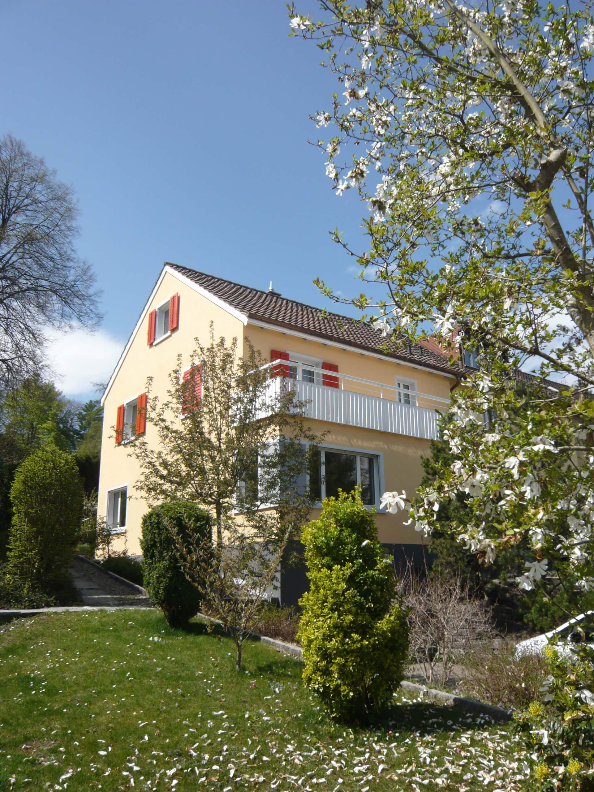 Wohnhaus B38 mit heller Fassade, roten Fensterläden und Garten.