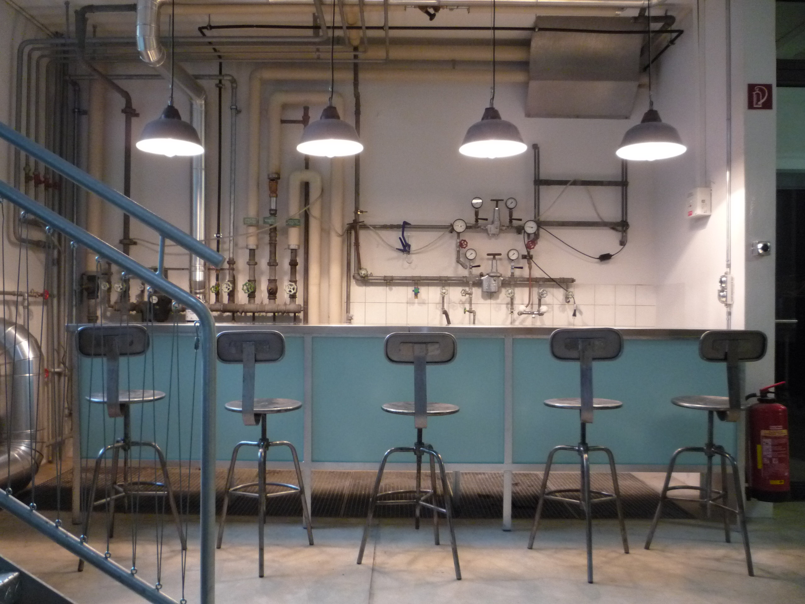 Bar mit türkisfarbener Front, Metallhockern und sichtbaren Installationsleitungen.