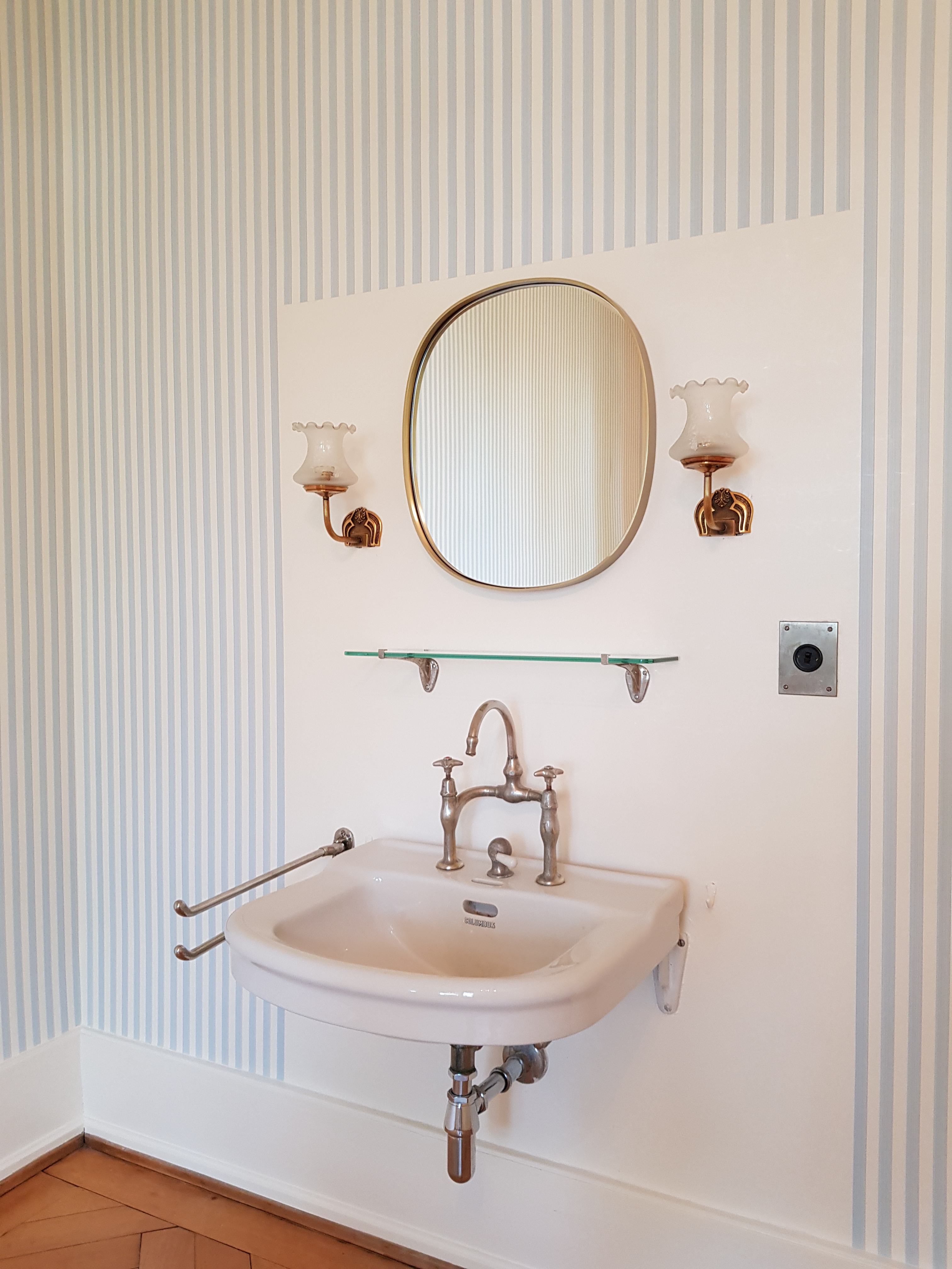 Waschbecken mit ovalem Spiegel und zwei Wandleuchten vor gestreifter Wand.