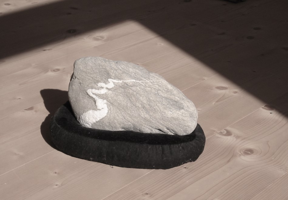 Flacher Naturstein auf dunklem Sockel im Sonnenlicht auf einem Holzboden.