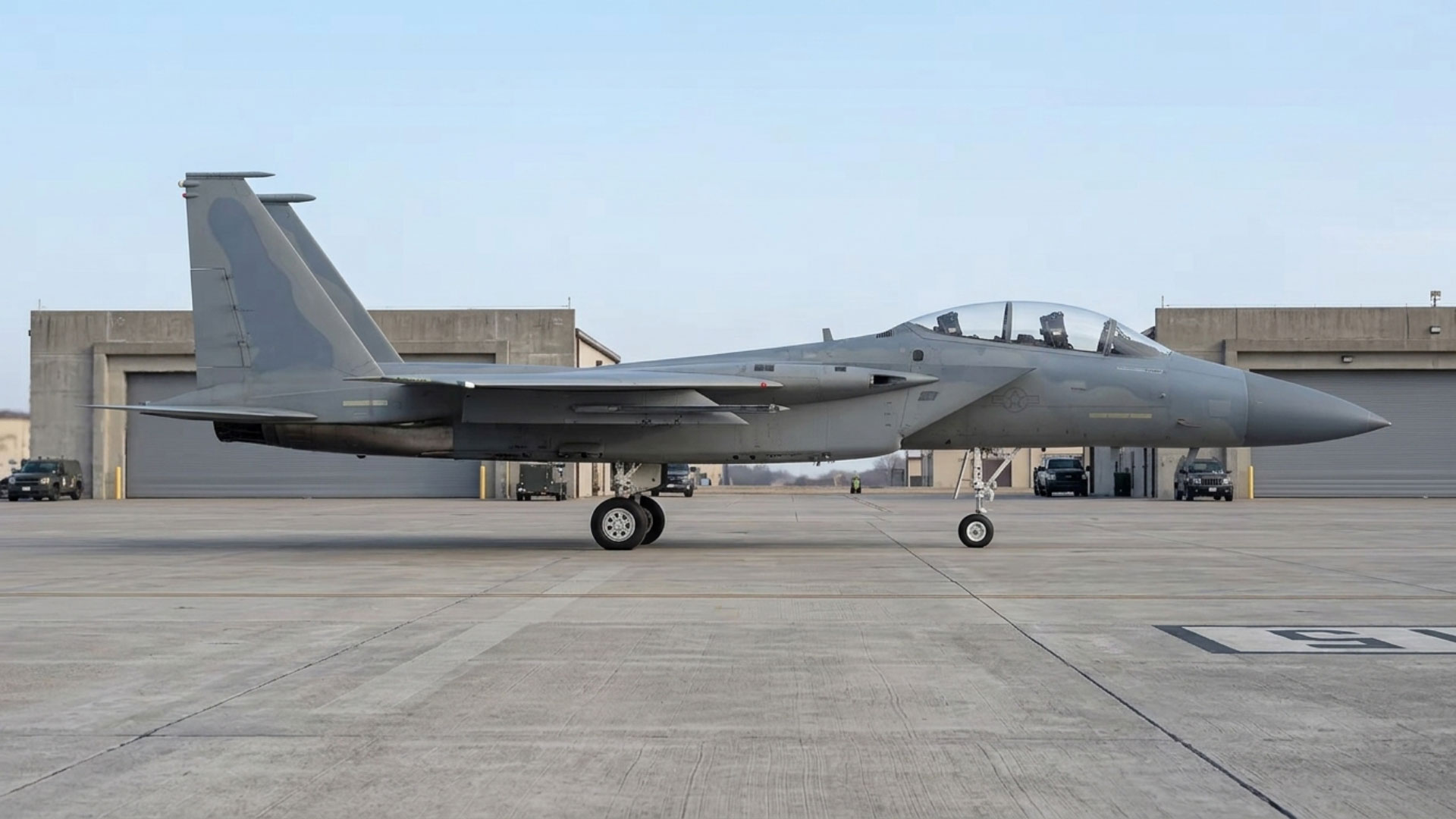 Grey twin-engine fighter jet parked on an airfield apron in front of hangars