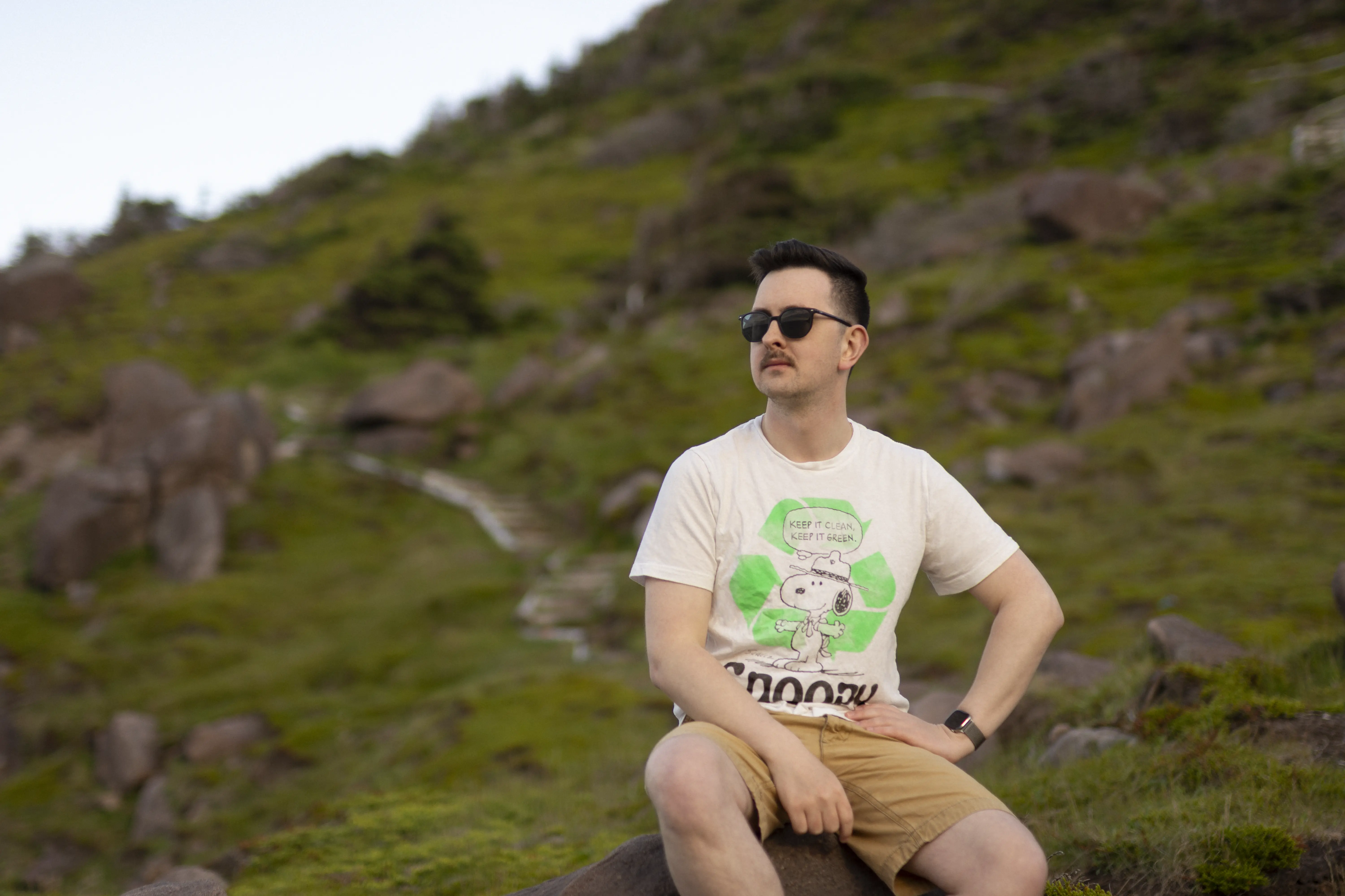 A picture of Cole sitting on a rock on the East Coast Trail. He is wearing sunglasses and a Snoopy shirt that says "Keep it clean. Keep it green." with a large neon green recycling symbol behind Snoopy.